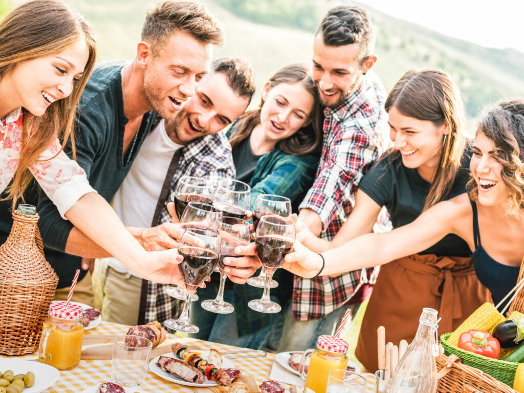 Young friends having genuine fun outdoor toasting red wine at open air bar-b-q party - Happy people eating grilled food in farmhouse vineyard winery - Youth friendship concept on warm contrast filter